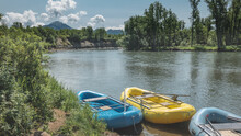 Inflatable Colorful Boats For Rafting Stand On The River Bank. There Is Lush Green Vegetation Around. A Mountain Range And A Volcano Are Visible Against A Background Of Blue Sky And Clouds. Kamchatka