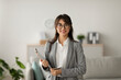 Portrait of cheerful female psychologist posing with clipboard, looking and smiling at camera, working in modern office