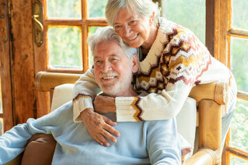 Wall Mural - Happy and enjoyed senior caucasian couple smile and hug each other sitting on a chair in cozy home cabin chalet - two man and woman old elderly people lifestyle enjoying relax and relationship