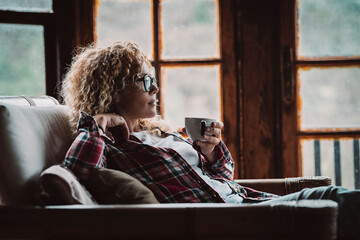 Wall Mural - Pretty adult hipster woman sitting and relaxing at home with cup of tea and windows in background - relaxed and enjoyed female people in cozy cabin house for holiday vacation or alternative rural