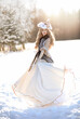 Artistic photo. A fantastic young blonde fairy in a historical costume and a hat with feathers stands in the cold wind. Winter nature background, white snow. The queen girl in a medieval dress.
