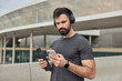 © WHstudio Leushin N - Muscular athletic man looks at smartphone screen downloads songs in playlist for training holds bottle with fresh water wears casual t shirt has regular workout poses outdoors. Healthy lifestyle