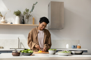 Happy pretty Black girl enjoying cooking hobby, chopping fresh vegetables for salad at kitchen table, making healthy dinner from organic bio ingredients, keeping vitamin rich nutrition