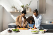 © fizkes - Happy African family couple teaching sweet daughter kid to cook healthy vegetarian dinner, chopping fresh vegetables for salad. Kin helping parents to prepare lunch at kitchen table