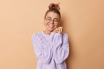 Wall Mural - Studio shot of happy young woman keeps hands near face smiles toothily has romantic tender facial expression recalls something pleasant in mind wears round spectacles and warm knitted sweater