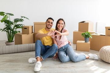 Wall Mural - Loving young international couple making heart with their hands, sitting on floor among boxes, moving to new house