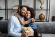 © fizkes - Cheerful cute Black daughter kid embracing grateful thankful dad holding surprise gift box due to birthday, fathers day. Happy father receiving present from child, hugging girl on sofa at home