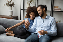 Happy Afro American Dad And Gen Z Teen Daughter Kid Using Smartphone At Home Together, Resting On Comfortable Sofa In Living Room, Making Video Call On Mobile Phone, Shopping Online, Browsing Internet