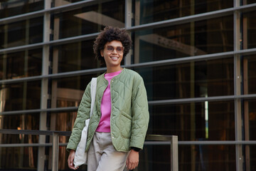 Wall Mural - Horizontal shot of happy woman with dark curly hair looks away joyfully wears trendy sunglasses and jacket carries fabric bag stands outdoors near building feels delighted. People and style concept