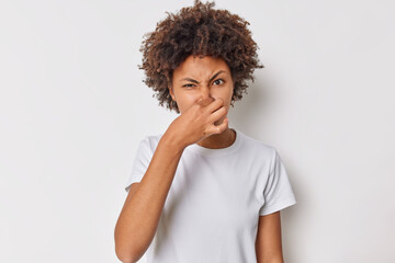 Wall Mural - Fie something stinks. Displeased curly haired woman covers nose smells something unpleasant has disgusting expression wears casual t shirt isolated over white background. Bad smell coming from kitchen