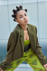 Wall Mural - Serious teenage girl with bun hairstyle looks directly at camera dressed in green stylish clothes wears bright makeup leans on knees poses against blurred background. People and style concept