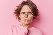Serious European woman tries to find decision thinks hard keeps finger near lips feels displeased has gloomy expression wears round spectacles long sleeved jumper isolated over pink background