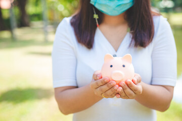 Wall Mural - money saving and business financial concept. business woman hand holding and putting money coin into piggy bank in the park outdoor against green blur nature background.