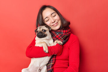 Happy young Asian woman embraces pug dog with love expresses love to favorite pet have friendly relationships enjoys company of best friend wears jumper with scarf isolated over red background