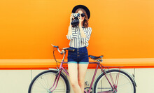 Summer Colorful Portrait Of Happy Smiling Young Woman Photographer Taking A Picture By Film Camera With Bicycle In The City On Orange Background