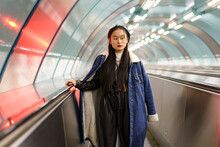 Portrait Of Stylish Asian Girl Wearing Trendy Street Fashion Outfit, Jeans Coat And Leather Beret On Escalator Riding Down To Underground Subway Station. Young Japanese Blogger Or Model In Urban Metro