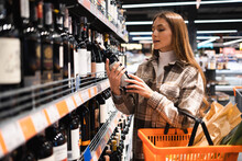 Beautiful Girl Carefully Chooses Wine In The Store. Woman Reads The Label On A Bottle Of Wine Before Buying