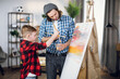 © sofiko14 - Handsome man and little boy in checkered shirts drawing abstract pattern on canvas. Happy father and son using brush and colorful paint during art process.