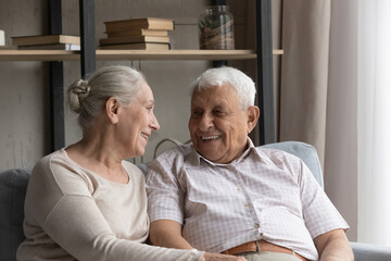 Wall Mural - Happy laughing candid older mature married family couple enjoying pleasant conversation, resting together on cozy couch. Loving smiling senior retired man and woman talking discussing life news.