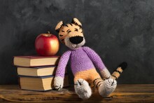 Handcrafted Toy Tiger Leaning Against A Stack Of Books With An Apple On Top Against A Gray Background.