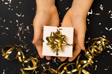 Child's hand gives gift box on black background with gold confetti . Composition for charity , christmas, new year , birthday, mother day, black friday , sale, promotions, xmas, or wedding. 