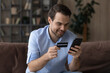 © fizkes - Smiling man holding smartphone and credit card, paying online, sitting on couch at home, satisfied customer looking at phone screen, making secure internet payment, shopping purchasing goods
