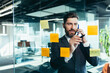 © Liubomir - businessman in reflection at a glass task board, a man talking on the phone, hanging plans on colored paper stickers, plans for the near future