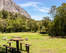 Wooden Table And Chairs In Grassy Picnic Area With Forest And Rocky Mountain In The Background, Tres Picos State Park, Teresopolis, Rio De Janeiro, Brazil