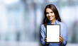 © vgstudio - Portrait of happy smiling brunette businesswoman showing mock up blank tablet pc, touchpad, blurred modern office interior background. Confident young business woman indoors.