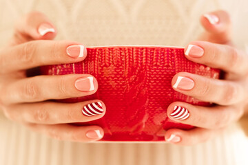 Woman's hands with french manicure and candy cane pattern on the nails. Woman with beautiful manicure holding big red knitted cup. The concept of cozy Christmas holidays and New Year.