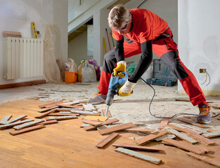 Parquet in wood glued to the slab, removed with an electric demolition hammer by a craftsman in workwear, in the apartment under renovation. In the foreground, the removed parquet slats. 