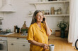 © shurkin_son - Beautiful grandmother in casual yellow clothes talking to her son on smartphone, looking aside, standing half-turned to camera near table counter in bright kitchen, having rest, spending leisure time