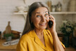 © shurkin_son - Close-up picture of attractive senior woman in yellow talking on phone to her friend in kitchen having surprised face expression, discussing funny things, smiling. Human emotions and feelings