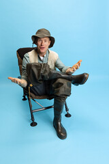 Poster - Portrait of young man, fisherman with fishing rod, spinning and equipment sitting isolated over blue studio background