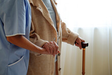 Senior Woman Holding Quad Cane Handle In Elderly Care Facility. Hospital Nurse Taking Care Of Mature Female With Walking Stick In Nursing Home. Background, Close Up On Hands With Wrinkled Skin.