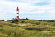 © Martin Piechotta - Tall red and white striped lighthouse building standing on small hill. Sandy coastal landscape with green vegetation and footpaths.