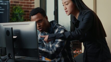 Two software developers coming at desk and sitting down holding laptop with coding interface and pointing at computer screen. Programmers team discussing algorithms looking at monitors with code.