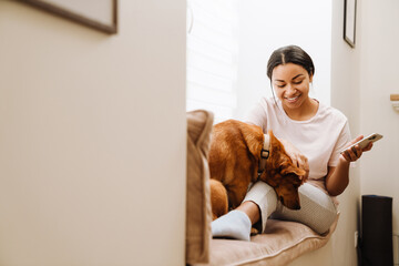 Wall Mural - Young hispanic woman petting her dog while using mobile phone