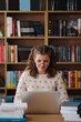 © arthurhidden - Attractive happy young girl student studying at the college library, sitting at the desk, using laptop computer, having video chat