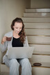 © DragonImages - Smiling young woman sitting on steps, drinking cup of morning coffee and reading article on laptop screen
