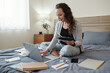 © DragonImages - Cheerful young female entrepreneur sitting on bed in her bedroom, eating tasty lunch and working on laptop