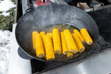 Close-up Heap Pile Of Many Sweet Tasty Ripe Boiled Corn Cobs Cooked In Cast Iron Cauldron On Open Fire Coal At Agricultural Food Fest Fair On Winter Day Outdoors. Takeaway Healthy Snack Food