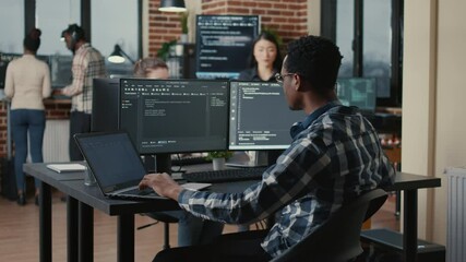 Portrait of system engineer fixing glasses while typing on laptop smiling satisfied about compiling results sitting at desk with multiple screens. App developer enjoying working on user interface.