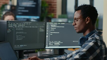 Sofware developer thinking while touching beard while typing on laptop sitting at desk with multiple screens parsing code. Focused database admin working with team coding in the background.
