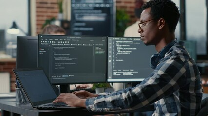 Portrait of african american developer using laptop to write code sitting at desk with multiple screens parsing algorithm in software agency. Coder working on user interface using portable computer.