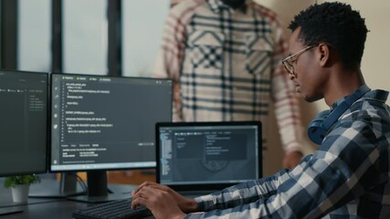 Focused software developer writing code on laptop looking at multiple screens with programming language is interrupted by colleague coder asking for advice. Programmers doing online cloud computing.