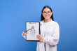 © tonefotografia - Young female ophthalmologist with glasses holding eye chart over blue background studio, healthcare concept