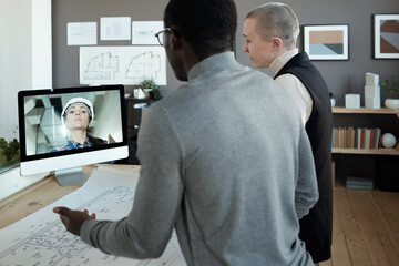 Canvas Print - Two contemporary architects communicating to female builder on computer screen while standing by table with blueprint