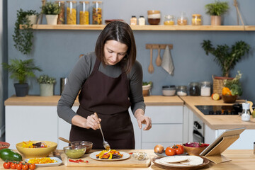 Wall Mural - Young female in apron flavoring vegetable food with homemade sauce pesto while bending by kitchen table and plate with cooked stew