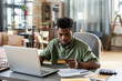 © pressmaster - Young African man looking at credit card in his hand while sitting by table in front of laptop and going to pay for online purchase